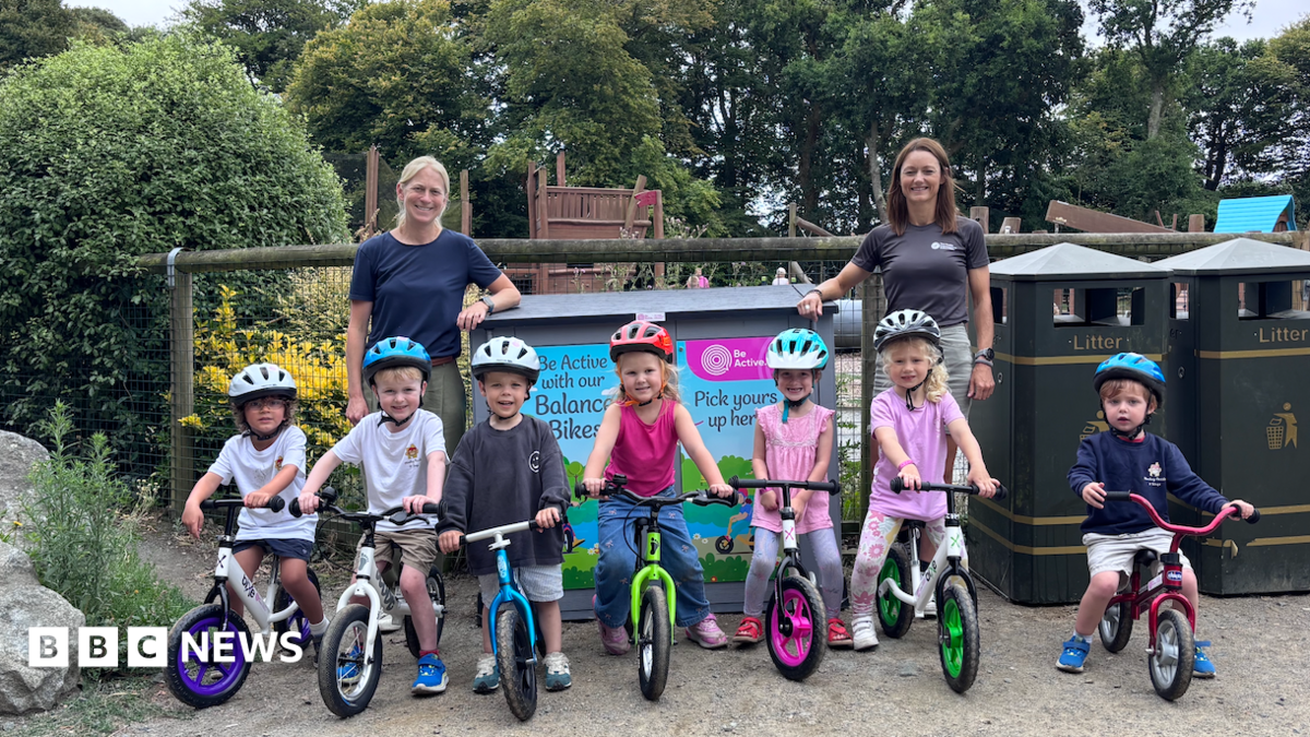 A row of children about age four and five on bikes, two women standing behind, at a playground. Trees and blue skies seen behind.