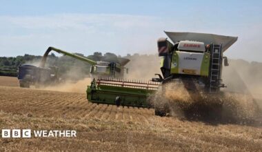 Farm machinery kicks up dust as it works on a brown, dry field, with trees in the distance and a milky blue sky overhead