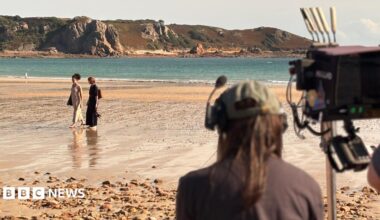 A camera man is seen from behind filminf two women walking along a sandy beach with cliffs in the background.