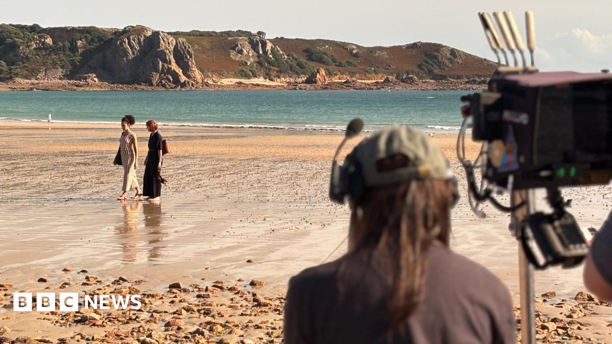 A camera man is seen from behind filminf two women walking along a sandy beach with cliffs in the background.