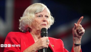 Ann Widdecombe speaking at the Reform UK party conference in 2024. She has a bob style haircut with mostly white hair, is wearing a red blazer with a broach, and is holding a microphone in her right hand as she speaks, while making a pointing gesture with her left hand.