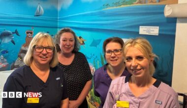 Four women standing in a hospital ward bathroom. Its walls are covered in a colourful mural which shows the sea, a beach, fish, dolphin, animals snorkelling and plants.