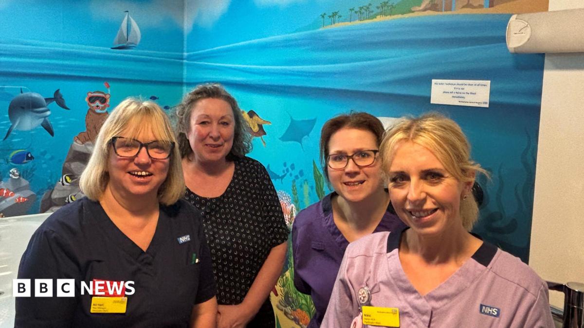 Four women standing in a hospital ward bathroom. Its walls are covered in a colourful mural which shows the sea, a beach, fish, dolphin, animals snorkelling and plants.