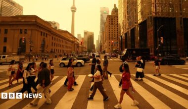 People walk on a crossing in a road downtown Toronto. There are buildings in the background and sky is orange from smoke.