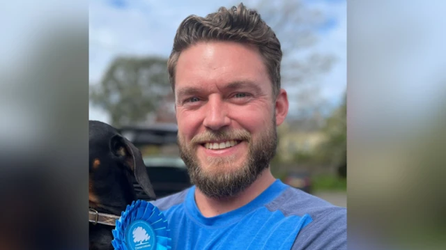 Phil Eckersley has greying blond hair and a beard. He is smiling while photographed wearing a blue t-shirt and a blue Conservative rosette.