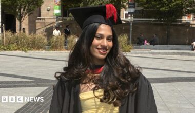 Person standing in a paved public square wearing a graduation gown and mortarboard with a tassel. A yellow dress is visible beneath the gown. Trees, ornamental grasses and stone buildings surround the square, with several people seated or walking in the background.