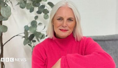 An woman with short white hair sits indoors on a grey cushioned seat, wearing a bright pink jumper and red trousers. A tall green houseplant in a woven basket stands behind them against a plain white wall.