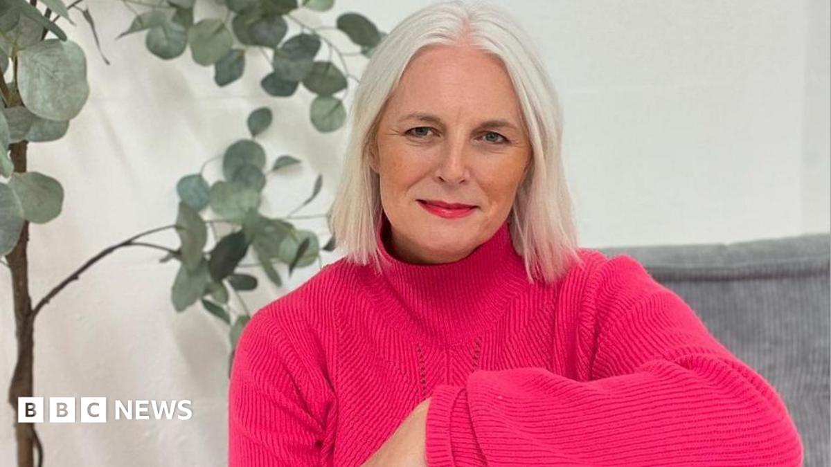 An woman with short white hair sits indoors on a grey cushioned seat, wearing a bright pink jumper and red trousers. A tall green houseplant in a woven basket stands behind them against a plain white wall.