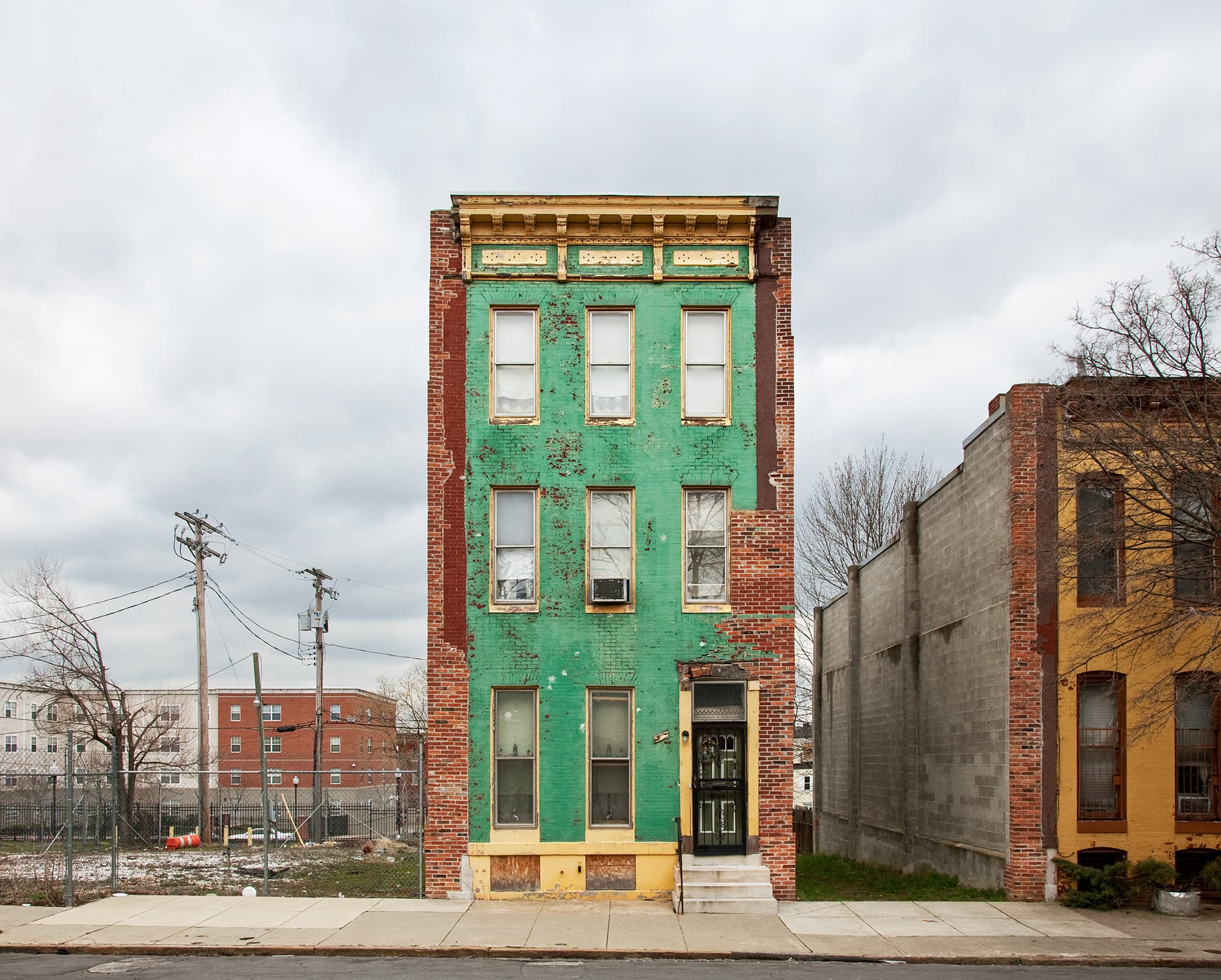 A solitary, weathered green brick rowhouse with peeled paint and a white staircase stands alone against a cloudy sky, separated from a neighboring yellow building by an empty gray concrete wall.