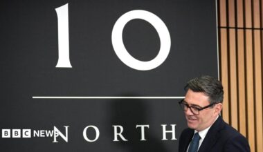 Andy Burnham holding a pint of beer at a bar, while looking across his shoulder to a person out of shot while talking. There is a camera crew and his new chancellor John Healey behind him. Burnham has short black hair and thick framed glasses and he is wearing a suit jacket over a white shirt. Healey, who has a bald head and is wearing a white shirt, is smiling.