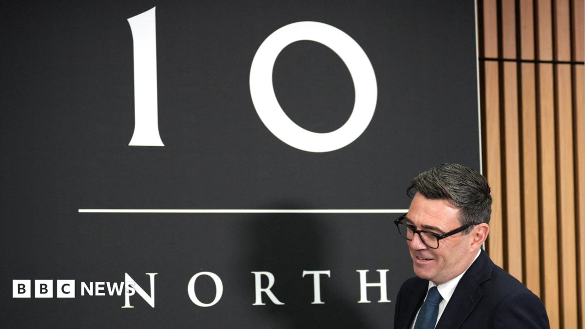 Andy Burnham holding a pint of beer at a bar, while looking across his shoulder to a person out of shot while talking. There is a camera crew and his new chancellor John Healey behind him. Burnham has short black hair and thick framed glasses and he is wearing a suit jacket over a white shirt. Healey, who has a bald head and is wearing a white shirt, is smiling.