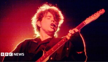 Tom Bailey plays a black electric guitar on a dark concert stage under intense red and orange lighting. Backlighting creates a bright halo around his hair during a live performance from the band's 1980s era at The Aragon Ballroom in Chicago, Illinois, on 27 April 1984.