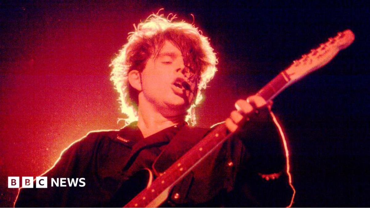 Tom Bailey plays a black electric guitar on a dark concert stage under intense red and orange lighting. Backlighting creates a bright halo around his hair during a live performance from the band's 1980s era at The Aragon Ballroom in Chicago, Illinois, on 27 April 1984.