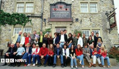 A large group of adults and children pose for a photograph outside a traditional stone-built pub called The Woolpack, with several people seated on hay bales in the front row. The building has a rustic stone façade, sash windows, climbing greenery, and prominent pub signs advertising ales, wine, and food. The group stretches across the entrance and frontage of the pub, creating the appearance of a cast or community gathering in a village setting.