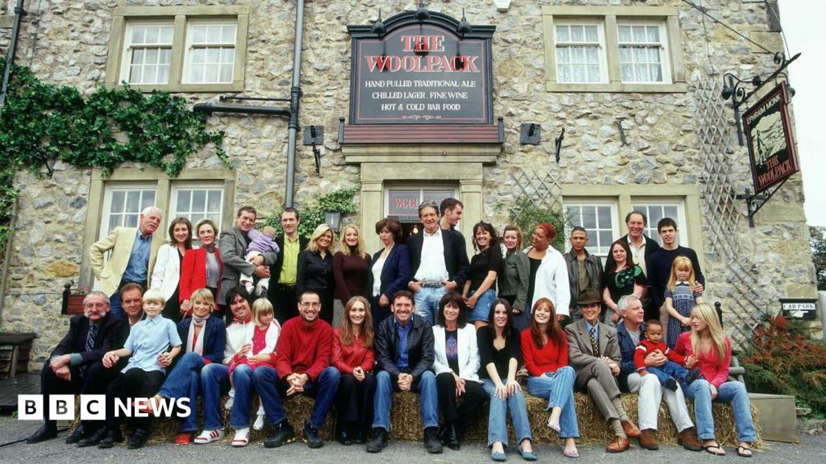 A large group of adults and children pose for a photograph outside a traditional stone-built pub called The Woolpack, with several people seated on hay bales in the front row. The building has a rustic stone façade, sash windows, climbing greenery, and prominent pub signs advertising ales, wine, and food. The group stretches across the entrance and frontage of the pub, creating the appearance of a cast or community gathering in a village setting.