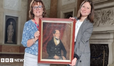 Two women hold a painting of a man in formal clothing. The women are stood in a grand room in a country house.