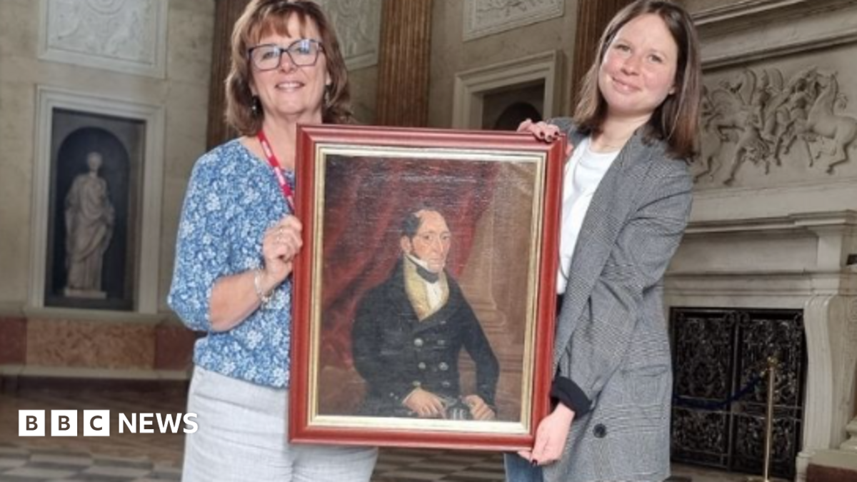 Two women hold a painting of a man in formal clothing. The women are stood in a grand room in a country house.