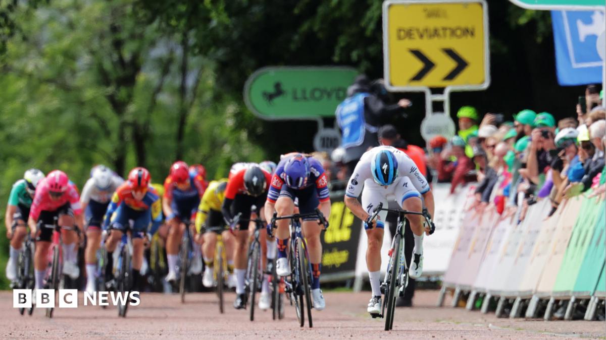 A tight knit group of about a dozen racing cyclists. To the left, spectators line the road behind a barrier and in the background, sightly out of focus, trees can be seen with signs saying "deviation" and "Lloyds" (the sponsors).