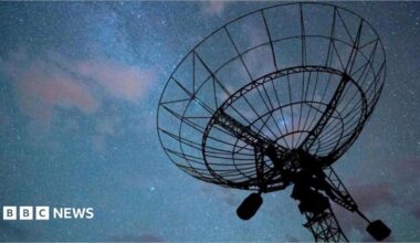 A radar disc pointed a dark night sky with an array of stars and clouds, tinged in purple, visible. The radar itself is in silhouette