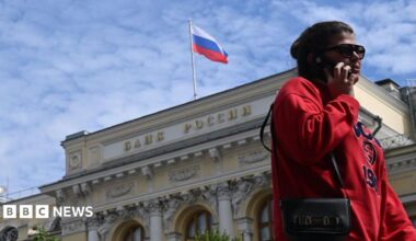 A woman wearing a red jumper and sunglasses, as well as a little bag on her right shoulder, talks on the phone in front of an exterior view of the Central Bank of the Russian Federation building on top of which the Russian flag can be seen