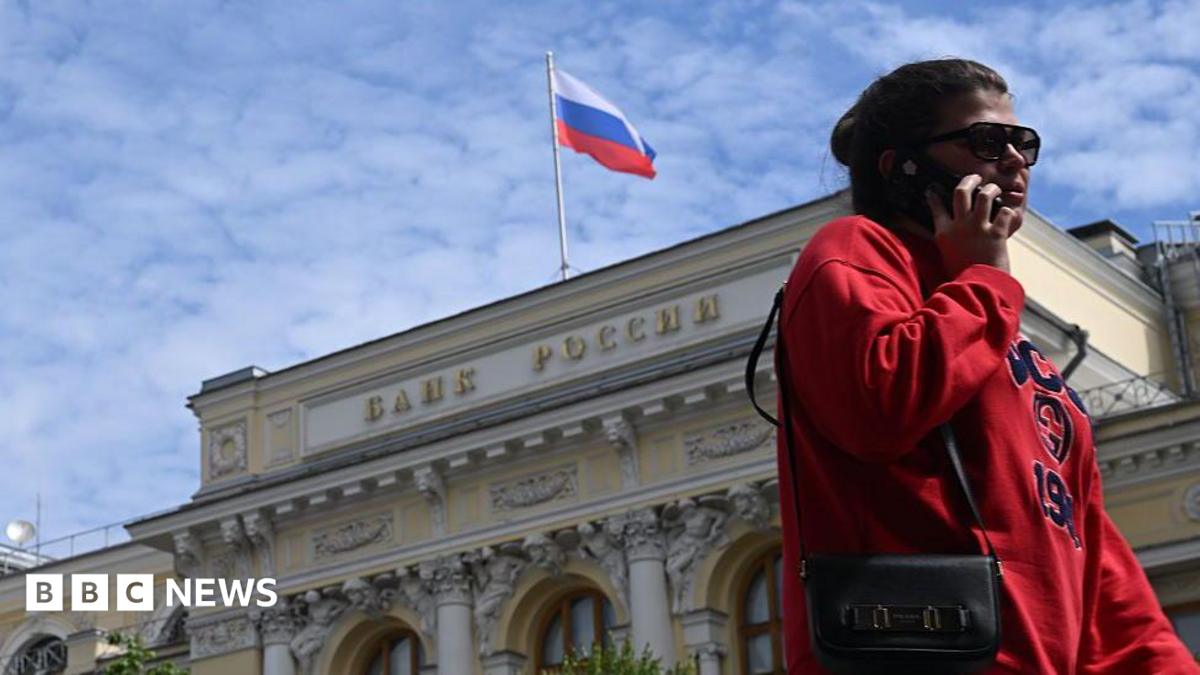 A woman wearing a red jumper and sunglasses, as well as a little bag on her right shoulder, talks on the phone in front of an exterior view of the Central Bank of the Russian Federation building on top of which the Russian flag can be seen