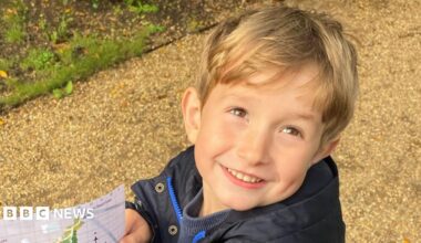 Benedict, a young boy with blond hair, on a path holding a map. He is wearing a black coat and is smiling to the camera.