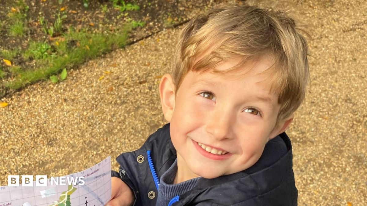 Benedict, a young boy with blond hair, on a path holding a map. He is wearing a black coat and is smiling to the camera.