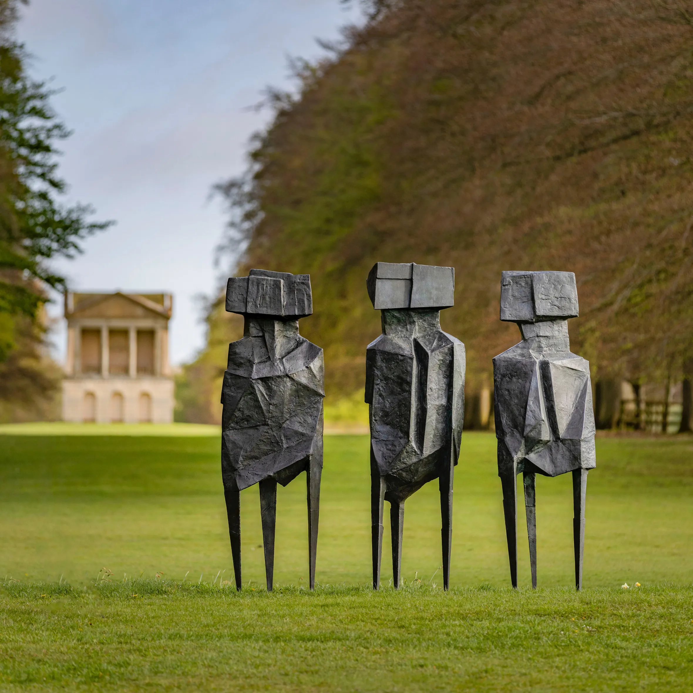 Three abstract, metallic sculptures of human-like figures standing in a grassy field with a classical building in the background.