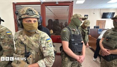 Men in military fatigues stand in a courtroom with a person sitting in a glass room