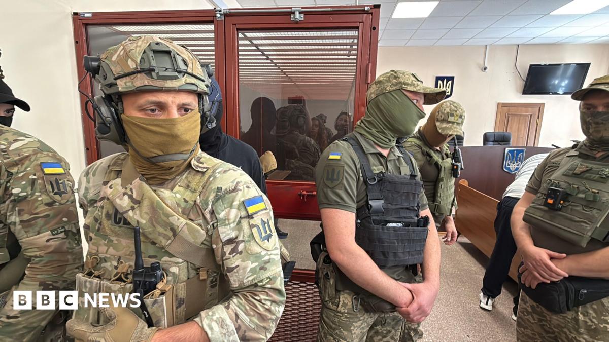 Men in military fatigues stand in a courtroom with a person sitting in a glass room