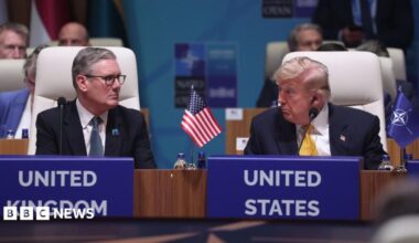 Sir Keir Starmer and Donald Trump are sat down in white chairs and looking at each other. There is a miniature United States flag between them on a wooden table and a miniature blue flag with the Nato emblem next to Trump's left arm on the table. Both men are wearing dark coloured suits, white shirts and ties.