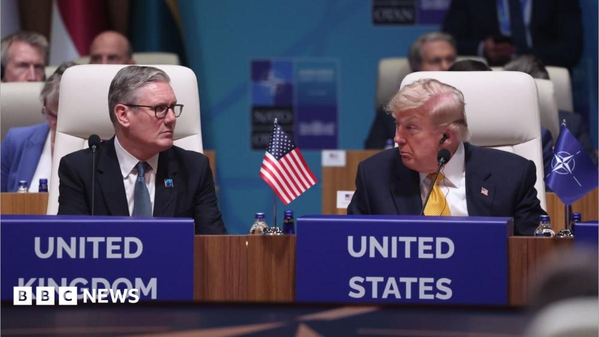 Sir Keir Starmer and Donald Trump are sat down in white chairs and looking at each other. There is a miniature United States flag between them on a wooden table and a miniature blue flag with the Nato emblem next to Trump's left arm on the table. Both men are wearing dark coloured suits, white shirts and ties.