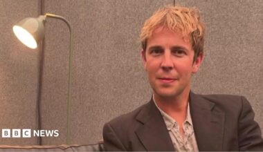 A head-and-shoulders image of Tom Odell sitting next to a lamp. He is smiling and looking into the camera.