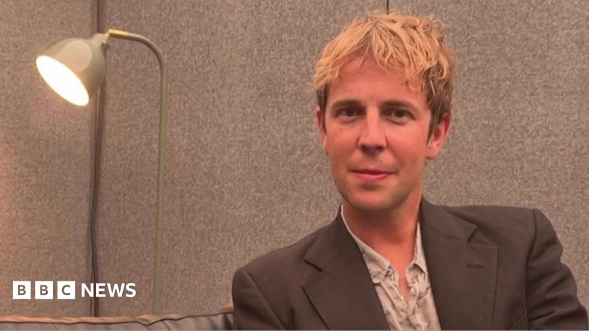 A head-and-shoulders image of Tom Odell sitting next to a lamp. He is smiling and looking into the camera.