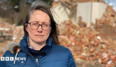 A woman in a blue puffy jacket looks at the camera. She has brown shoulder length hair and is wearing glasses. Behind her is a pile of red brick rubble and a partly demolished house.
