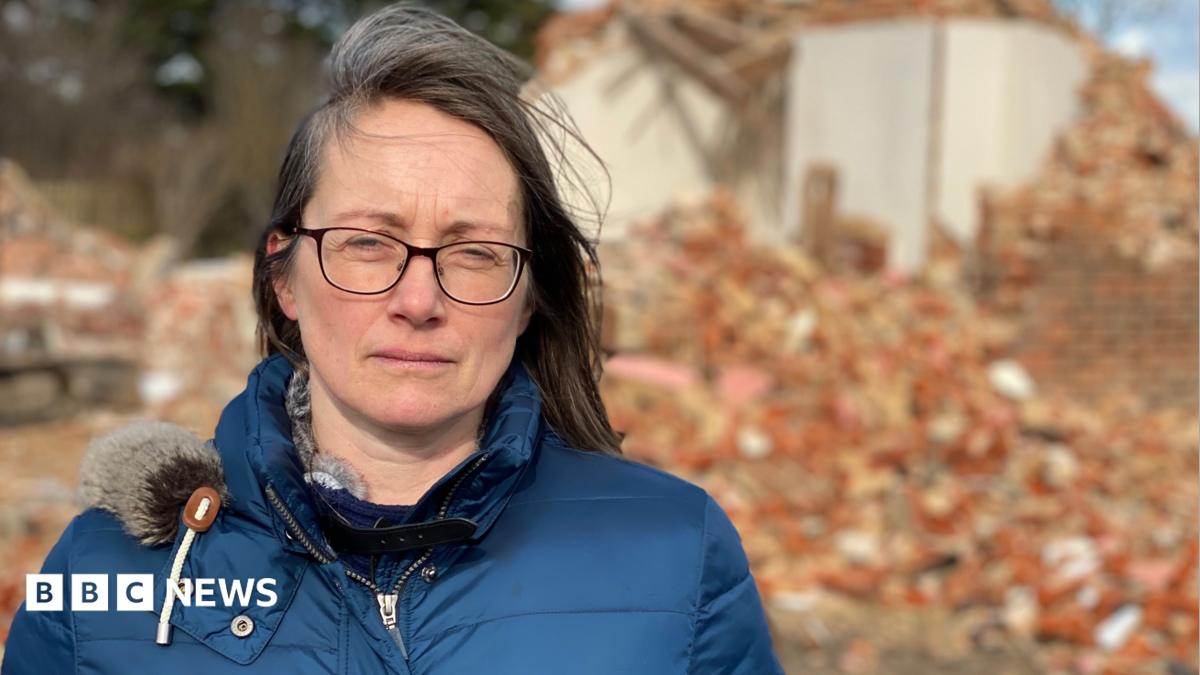 A woman in a blue puffy jacket looks at the camera. She has brown shoulder length hair and is wearing glasses. Behind her is a pile of red brick rubble and a partly demolished house.