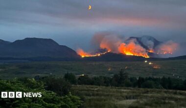 A line of fire on a mountain set against a darkening sky. Fields can be seen in the foreground, with the moon visible in the night sky.
