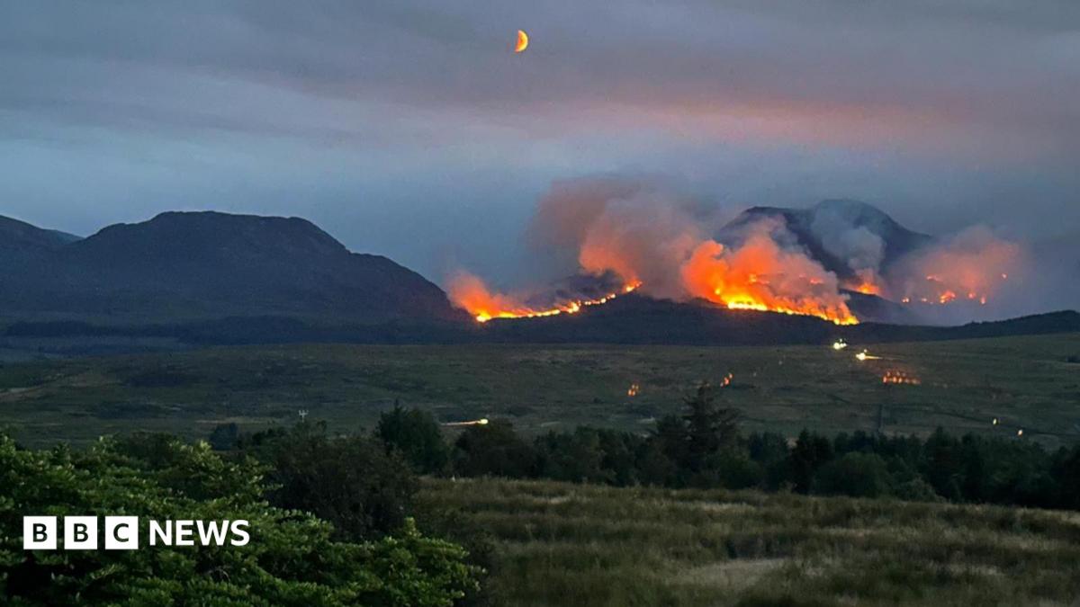 A line of fire on a mountain set against a darkening sky. Fields can be seen in the foreground, with the moon visible in the night sky.