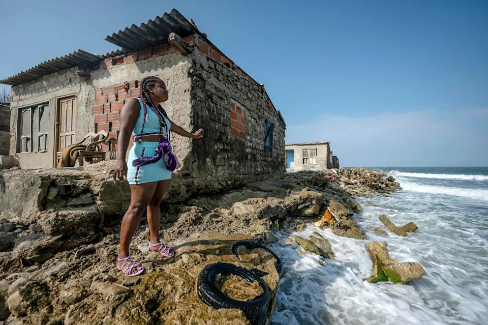 A woman stands outside an old home showing where sea level rise has eroded the shoreline nearly to the home's foundation.