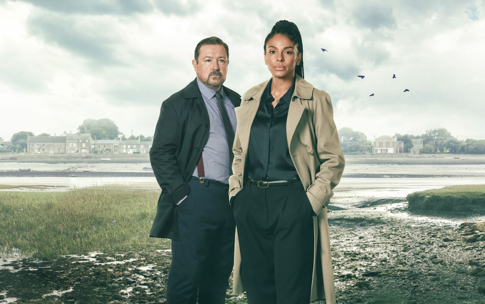 The Bay two individuals in trench coats standing on a marshy landscape with a cloudy sky and houses in the background.