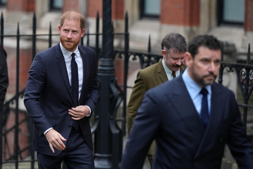 Prince Harry walks in a navy suit outside on the street behind several men.
