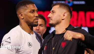 Anthony Joshua and Kristian Prenga face off as they look each other in the eye, Joshua wearing a white top and Prenga in a black hoodie