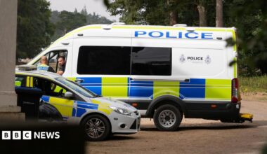 A large police van and a police car parked next to each other. Two officers - including one in the passenger seat and one standing outside - talking