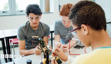 Three students wearing safety goggles work together on a robotic arm in a classroom or laboratory, connecting wires and examining components at a workbench. Tables, chairs and large windows are visible in the bright background.