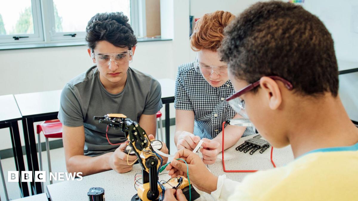 Three students wearing safety goggles work together on a robotic arm in a classroom or laboratory, connecting wires and examining components at a workbench. Tables, chairs and large windows are visible in the bright background.