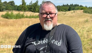 A man with short grey hair, a white goatee and black-rimmed glasses standing on a patch of grassland on a sunny day. Many trees are in the distance. He is wearing a black Motorhead t-shirt and has a pair of binoculars hanging around his neck.