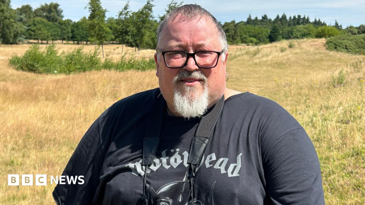 A man with short grey hair, a white goatee and black-rimmed glasses standing on a patch of grassland on a sunny day. Many trees are in the distance. He is wearing a black Motorhead t-shirt and has a pair of binoculars hanging around his neck.