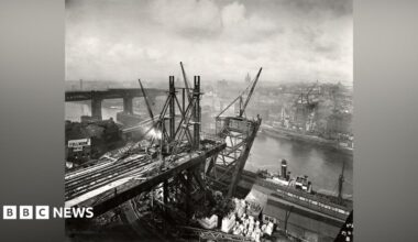 A black and white photograph showing the building on the Tyne Bridge from the Gateshead side. Construction is taking place on either side of the river, but the arch going over the water is just beginning to take shape. There are factories and buildings surrounding the structure, with the High Level Bridge and Swing Bridge visible behind it to the left. There are multiple cranes working on the bridge. The sky is covered in white clouds.