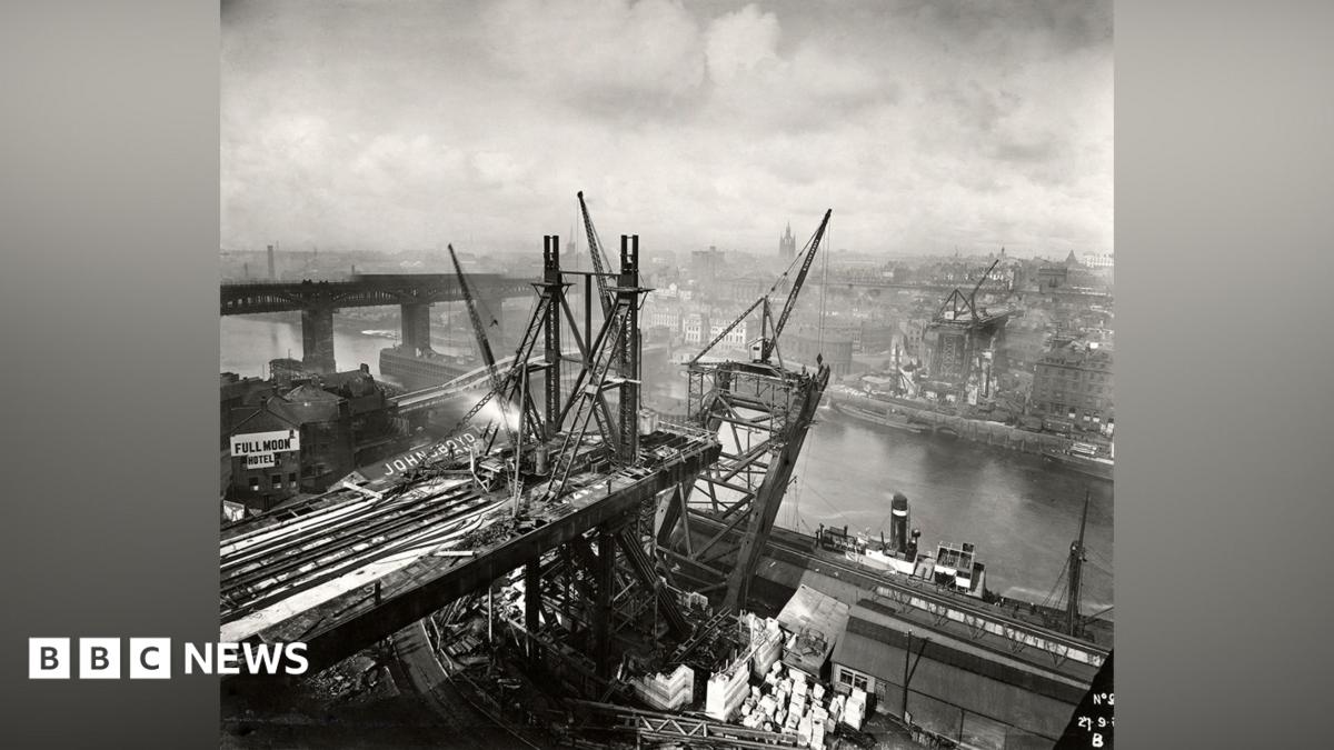 A black and white photograph showing the building on the Tyne Bridge from the Gateshead side. Construction is taking place on either side of the river, but the arch going over the water is just beginning to take shape. There are factories and buildings surrounding the structure, with the High Level Bridge and Swing Bridge visible behind it to the left. There are multiple cranes working on the bridge. The sky is covered in white clouds.