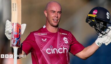 Chris Lynn, wearing Northamptonshire's red shirt, raises his bat and helmet to celebrate scoring a century against Glamorgan